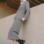A stylish woman wearing a sophisticated grey pinstripe high-waisted midi skirt with a front slit, paired with a white button-up shirt rolled at the sleeves and black ankle boots for a minimalist corporate chic look.