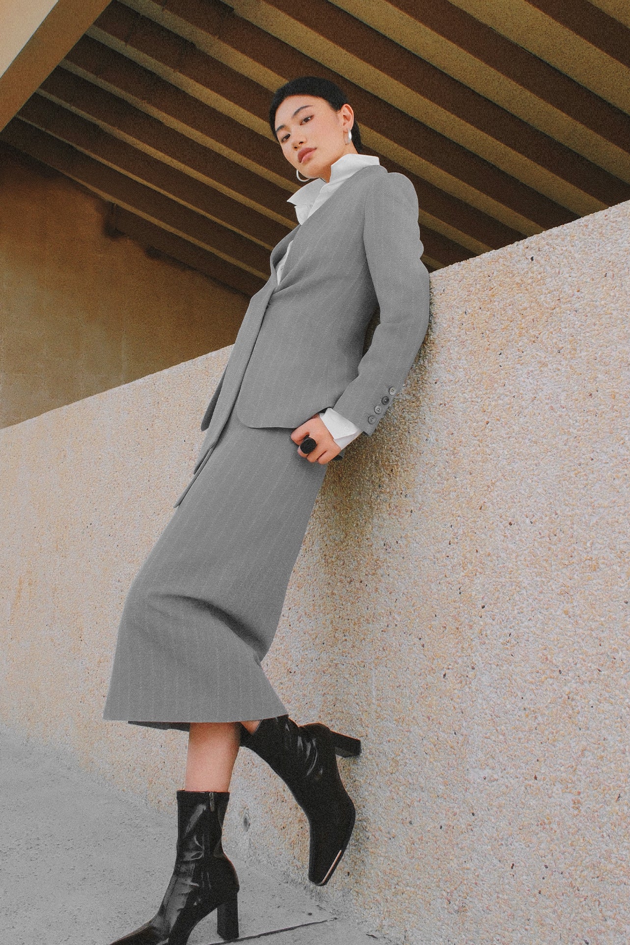 A stylish woman wearing a sophisticated grey pinstripe high-waisted midi skirt with a front slit, paired with a white button-up shirt rolled at the sleeves and black ankle boots for a minimalist corporate chic look.