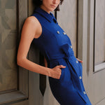 An elegant woman wearing a sophisticated royal cobalt blue trench dress featuring a structured turn-down collar, a cropped capelet overlay, a double-breasted button front, and a cinched self-tie belted waist.