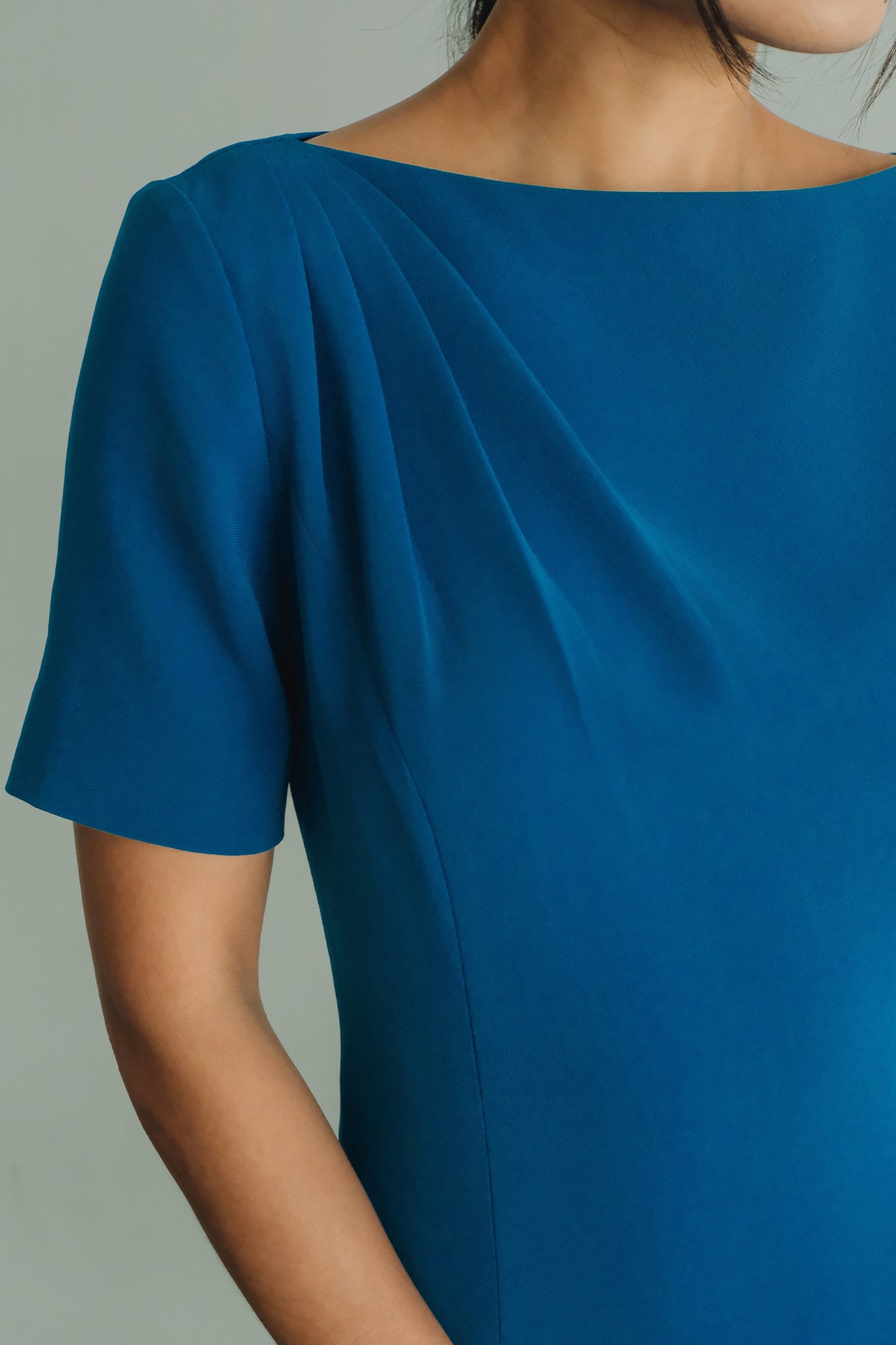 A beautiful model wearing a sophisticated medium blue A-line midi dress featuring an elegant boat neckline with refined asymmetric shoulder pleating, short sleeves, and a flowing flared swing skirt.