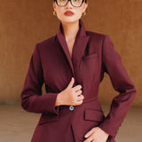 A professional woman wearing glasses and an elegant deep burgundy skirt suit featuring a sculptural stand-up lapel blazer with a single-button closure, tailored flap pockets, and a matching high-waisted pencil skirt.
