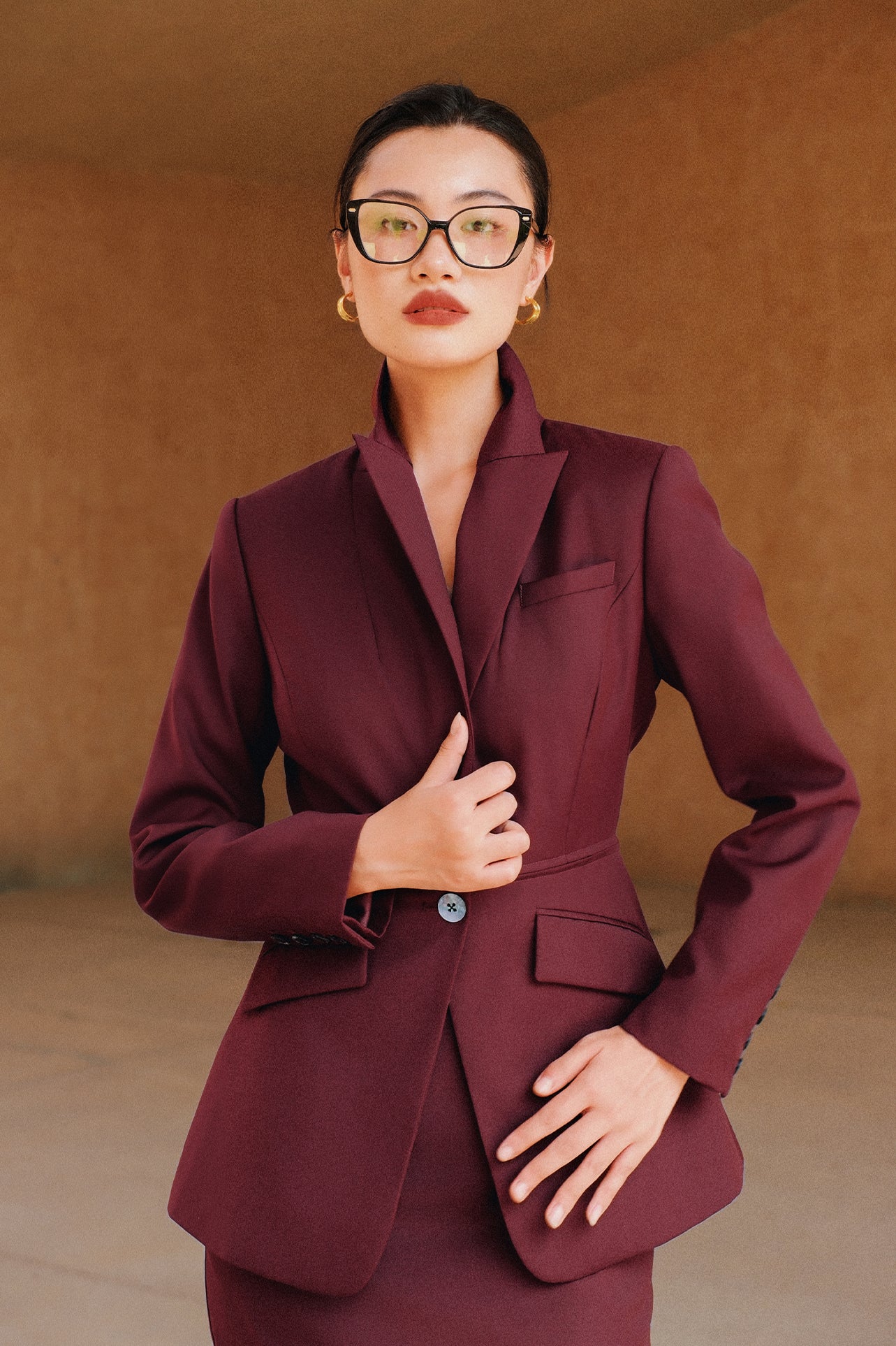 A professional woman wearing glasses and an elegant deep burgundy skirt suit featuring a sculptural stand-up lapel blazer with a single-button closure, tailored flap pockets, and a matching high-waisted pencil skirt.