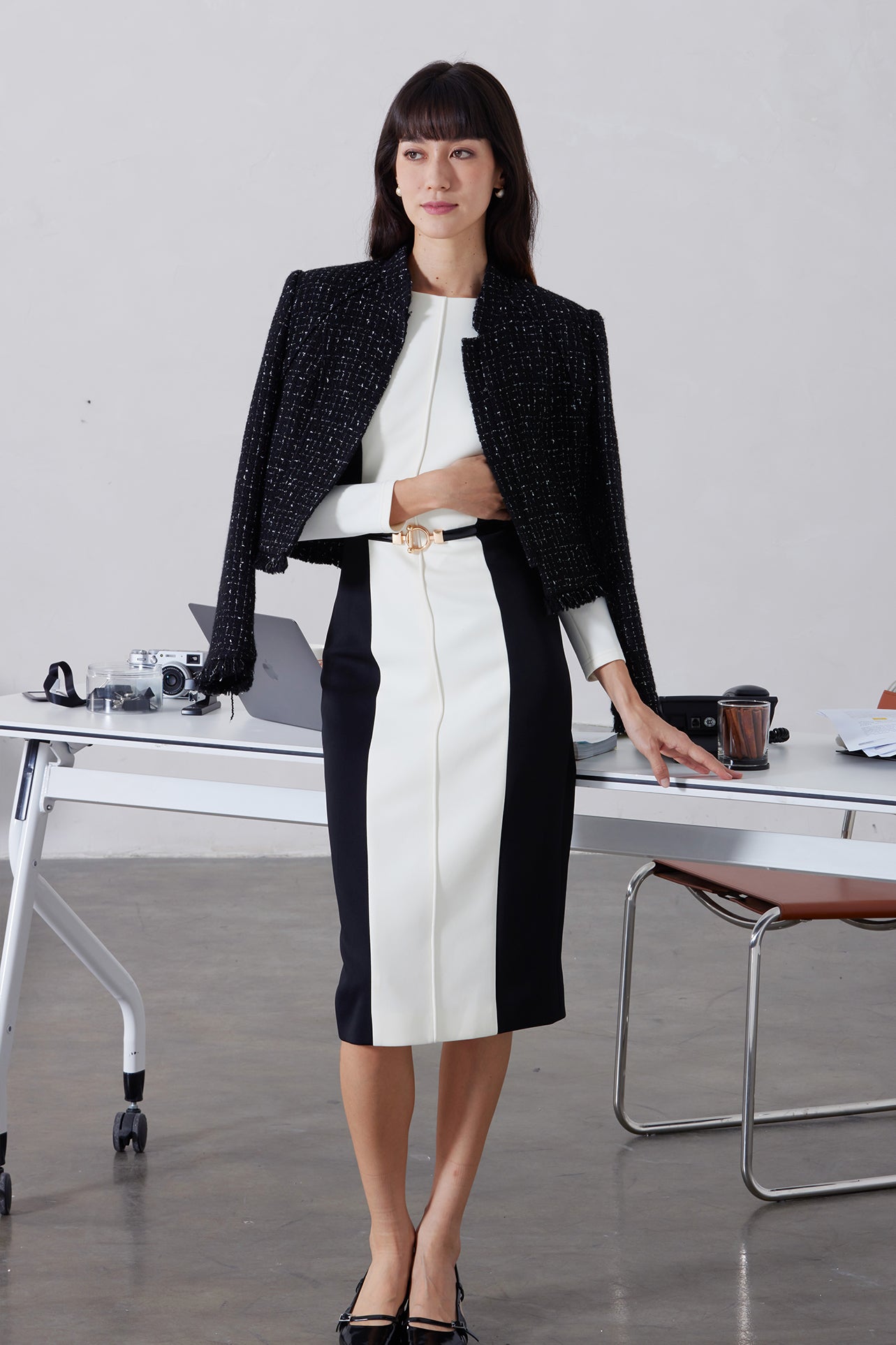 A professional woman wearing a sophisticated black and white color-block midi dress, featuring slimming side panels, long sleeves, and an elegant black belt with a gold buckle for a modern executive office look.
