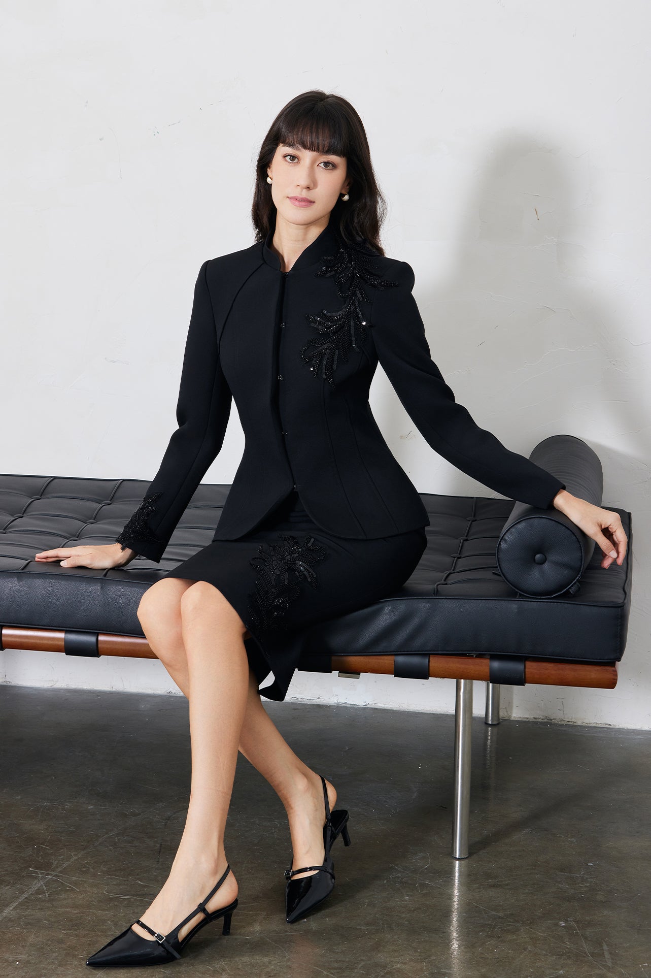A professional woman wearing a sophisticated black skirt suit, featuring a mandarin collar with elegant leaf embroidery on the shoulder and skirt, and traditional frog button detailing for a modern oriental executive look.