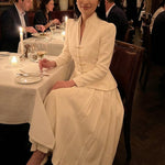 A sophisticated woman enjoying coffee in Paris wearing an elegant cream white modern qipao suit, featuring a mandarin collar blazer with traditional frog buttons and a matching pleated skirt for a quiet luxury oriental look.