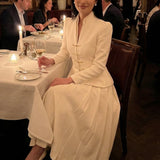 A sophisticated woman enjoying coffee in Paris wearing an elegant cream white modern qipao suit, featuring a mandarin collar blazer with traditional frog buttons and a matching pleated skirt for a quiet luxury oriental look.