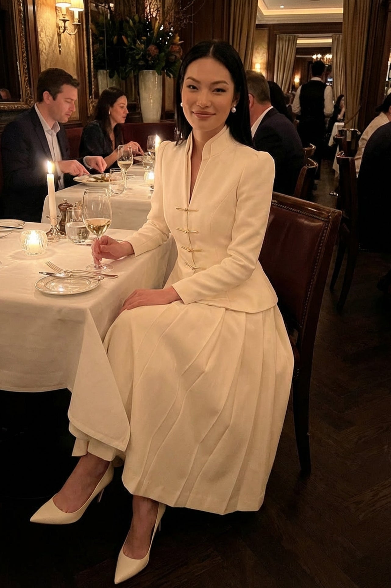 A sophisticated woman enjoying coffee in Paris wearing an elegant cream white modern qipao suit, featuring a mandarin collar blazer with traditional frog buttons and a matching pleated skirt for a quiet luxury oriental look.
