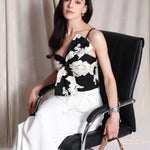 A professional woman wearing a sophisticated one-shoulder black and white floral print top, featuring a dramatic oversized bow detail on the shoulder, paired with white high-waisted trousers for an elegant evening look.