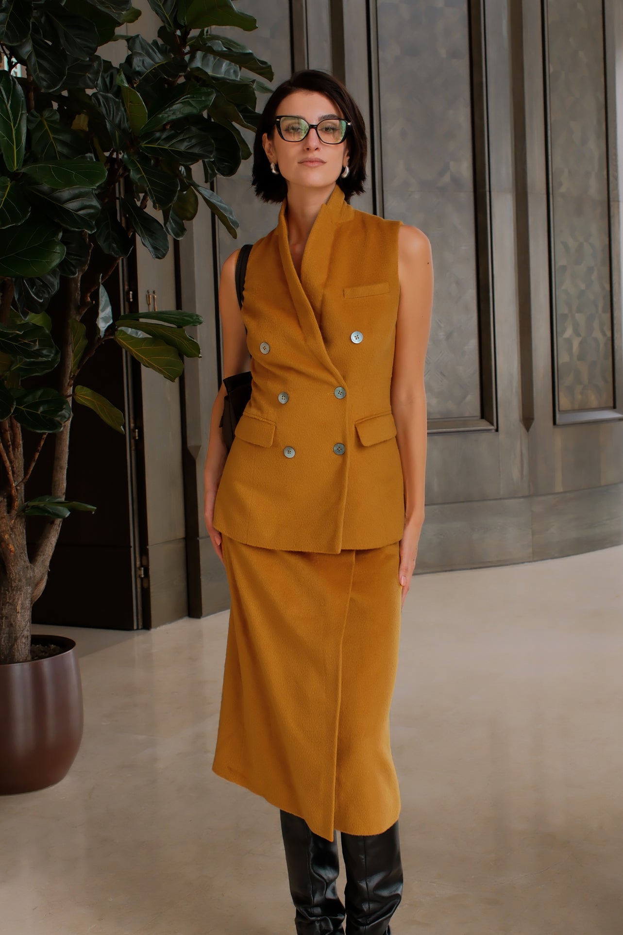 An elegant Asian woman posing in a luxurious camel wool two-piece suit featuring a sleeveless double-breasted longline vest cinched with a wide black belt, paired with a matching midi column skirt and black opera gloves.