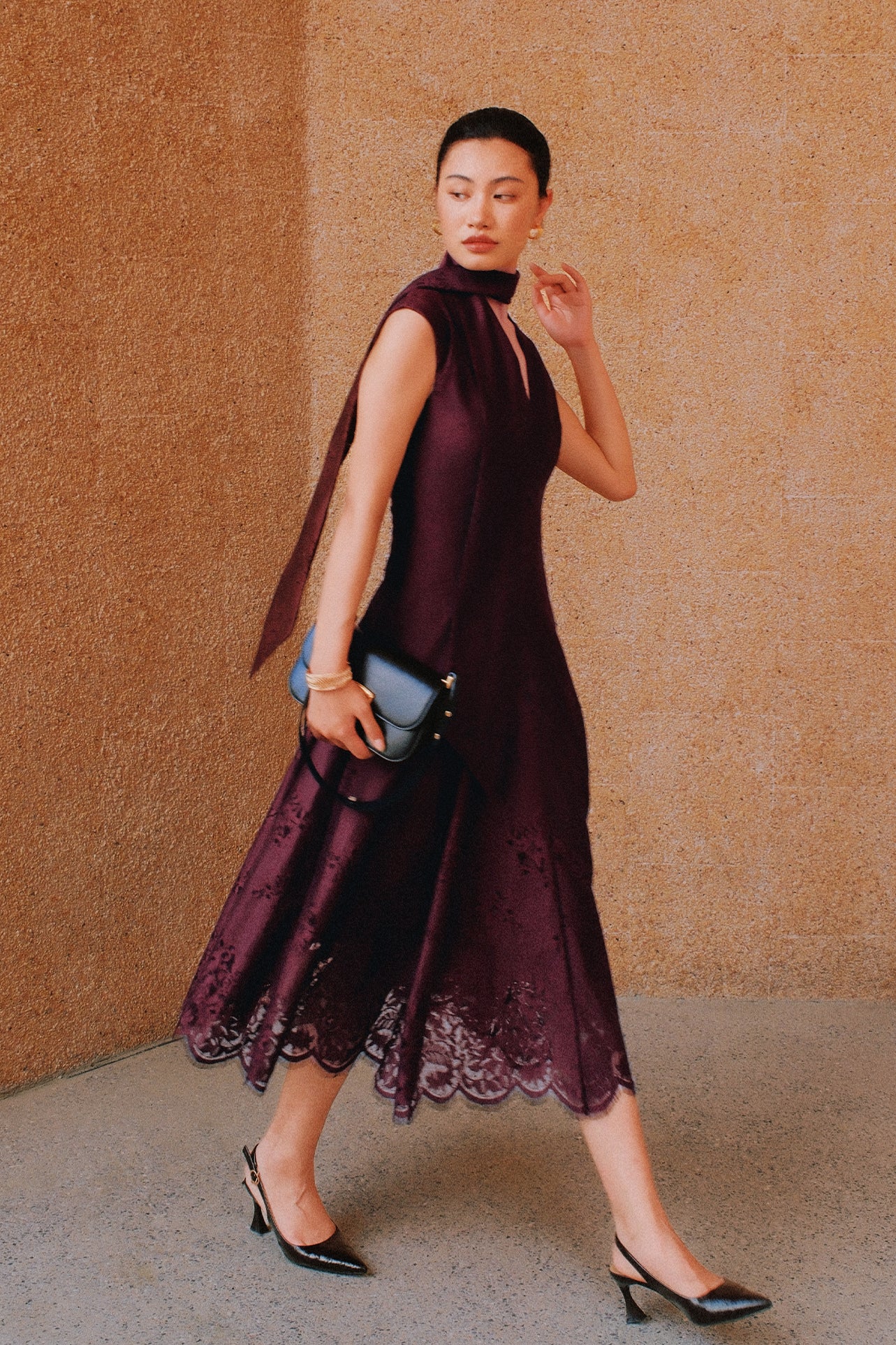 A chic woman wearing sunglasses and an elegant deep burgundy sleeveless V-neck A-line midi dress featuring textured jacquard fabric, delicate lace hemline, and a matching flowing neck scarf.