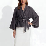 Two professional models wearing elegant shimmering satin wrap tops with kimono-style sleeves and waist ties, one in champagne gold and the other in charcoal grey with contrast white piping, paired with ivory high-waisted trousers.