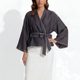 Two professional models wearing elegant shimmering satin wrap tops with kimono-style sleeves and waist ties, one in champagne gold and the other in charcoal grey with contrast white piping, paired with ivory high-waisted trousers.