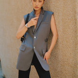 A style comparison showing two ways to wear a longline sleeveless blazer vest: on the left, a slate blue vest layered over a white shirt and cinched with a brown belt worn as a dress with knee-high boots; on the right, an oatmeal beige vest styled with flowing white wide-leg trousers.