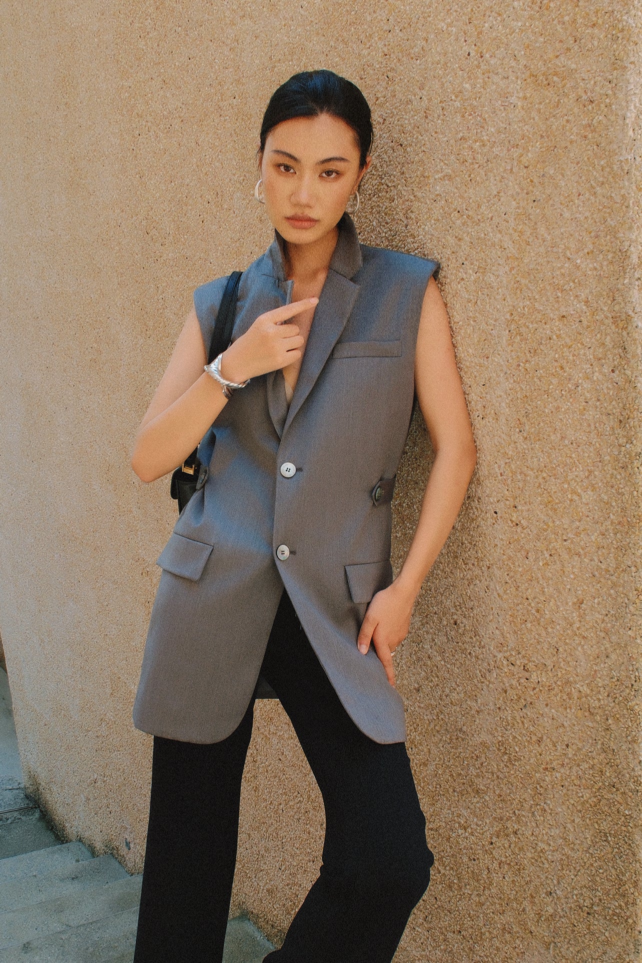 A style comparison showing two ways to wear a longline sleeveless blazer vest: on the left, a slate blue vest layered over a white shirt and cinched with a brown belt worn as a dress with knee-high boots; on the right, an oatmeal beige vest styled with flowing white wide-leg trousers.