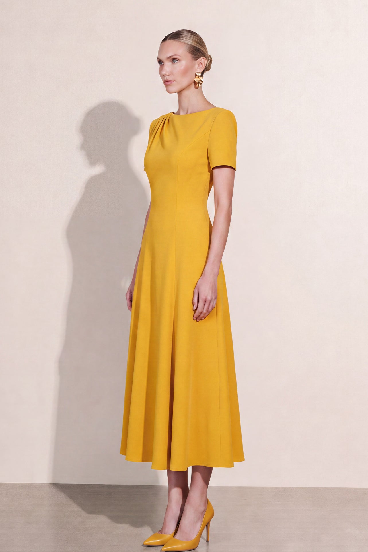 A blonde model wearing a sophisticated mustard yellow A-line midi dress featuring an elegant boat neckline with subtle shoulder pleating, short sleeves, and a flattering flared skirt.