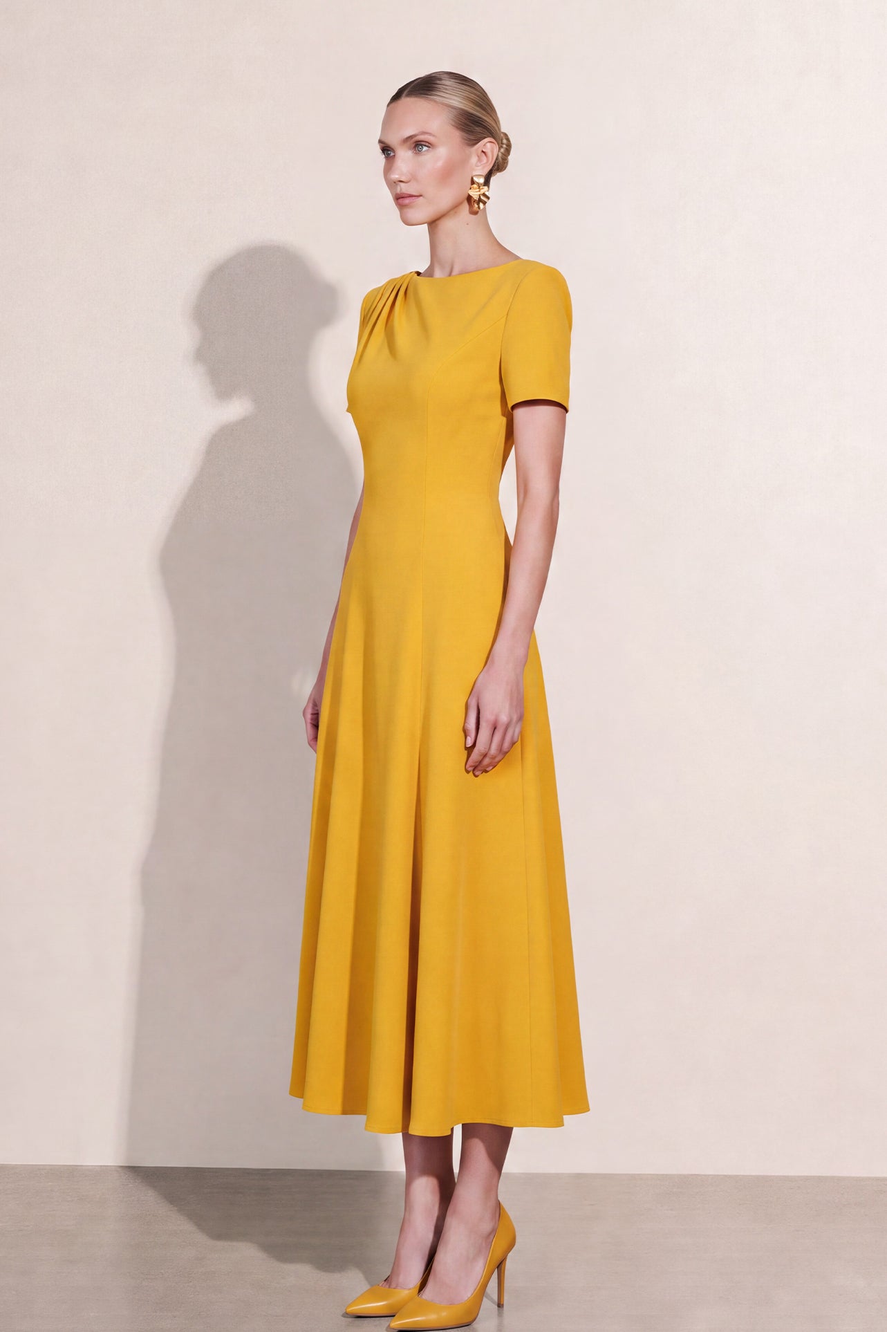 A blonde model wearing a sophisticated mustard yellow A-line midi dress featuring an elegant boat neckline with subtle shoulder pleating, short sleeves, and a flattering flared skirt.