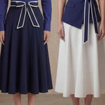 A comparison of two elegant professional looks: a navy blue pleated midi skirt and a crisp white flare skirt, both featuring sophisticated contrast-piped bow belts for a polished executive appearance.