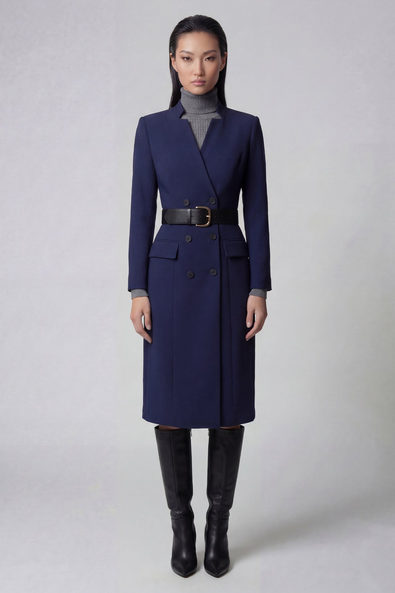 An elegant professional woman wearing a sophisticated navy blue double-breasted long coat dress, featuring a black waist belt and structured shoulders for a powerful executive look.