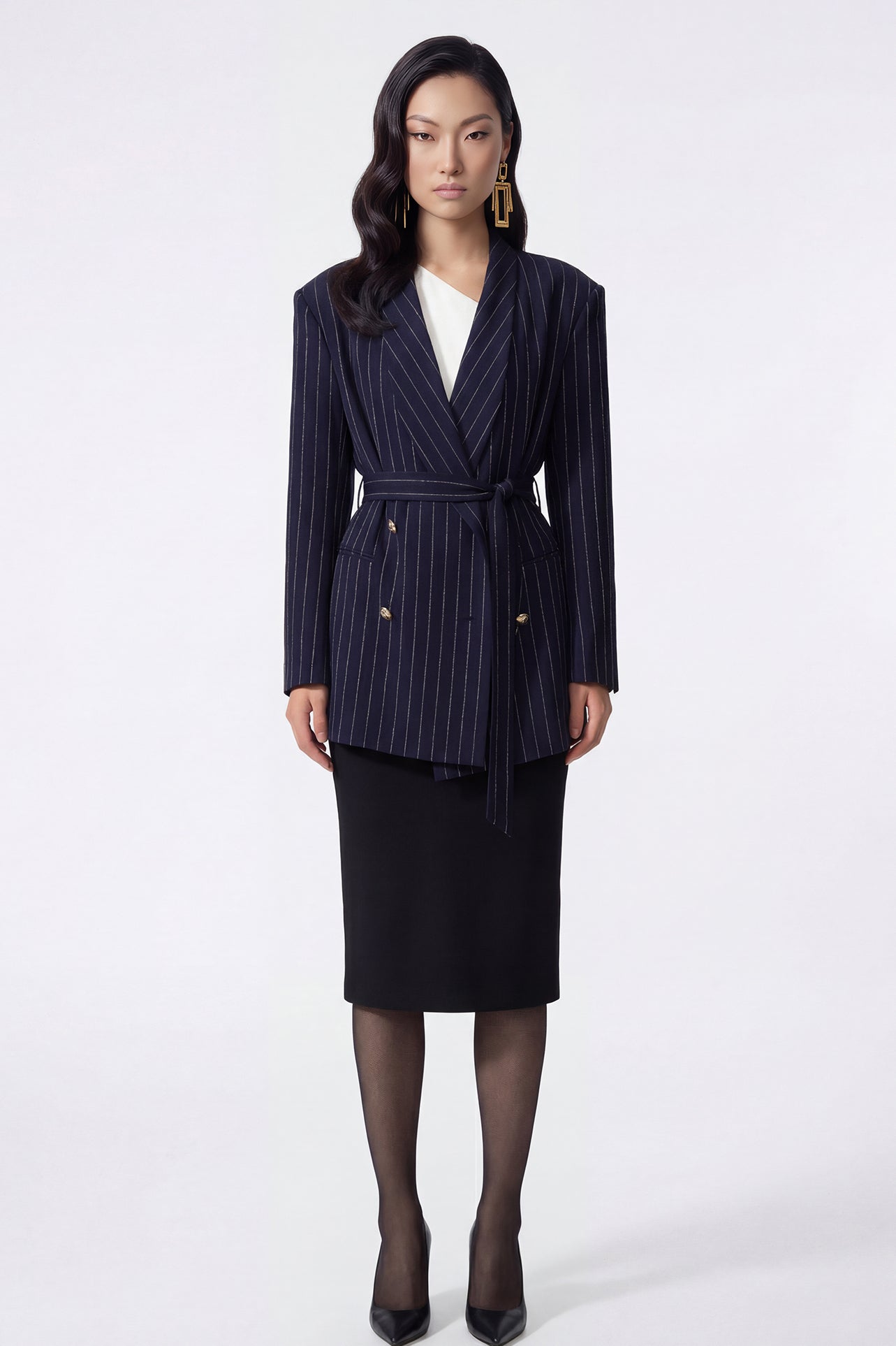 A professional woman wearing a sophisticated navy blue pinstripe blazer jacket, featuring a longline silhouette with a matching tie-waist belt and gold earring accents for an elegant executive office look.