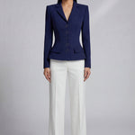 A professional model wearing a sophisticated navy blue tailored blazer with an hourglass silhouette and unique O-ring gold buttons, paired with crisp white trousers for an executive office look.