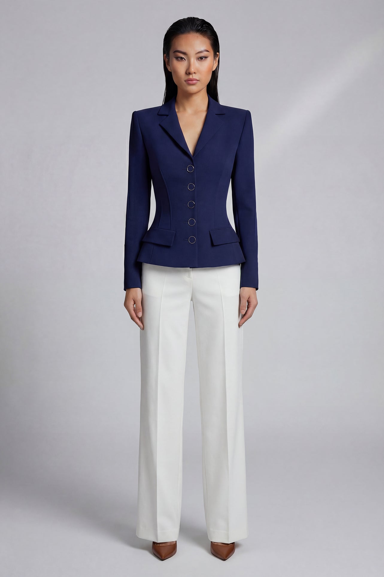 A professional model wearing a sophisticated navy blue tailored blazer with an hourglass silhouette and unique O-ring gold buttons, paired with crisp white trousers for an executive office look.