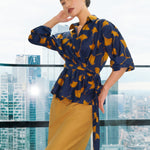 A stylish woman posing against a city skyline wearing a navy blue wrap blouse with golden ginkgo leaf print tied at the waist, paired with tailored mustard yellow wide-leg trousers.