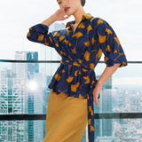 A stylish woman posing against a city skyline wearing a navy blue wrap blouse with golden ginkgo leaf print tied at the waist, paired with tailored mustard yellow wide-leg trousers.