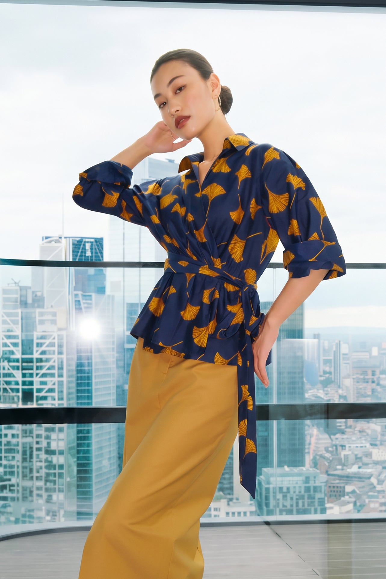 A stylish woman posing against a city skyline wearing a navy blue wrap blouse with golden ginkgo leaf print tied at the waist, paired with tailored mustard yellow wide-leg trousers.