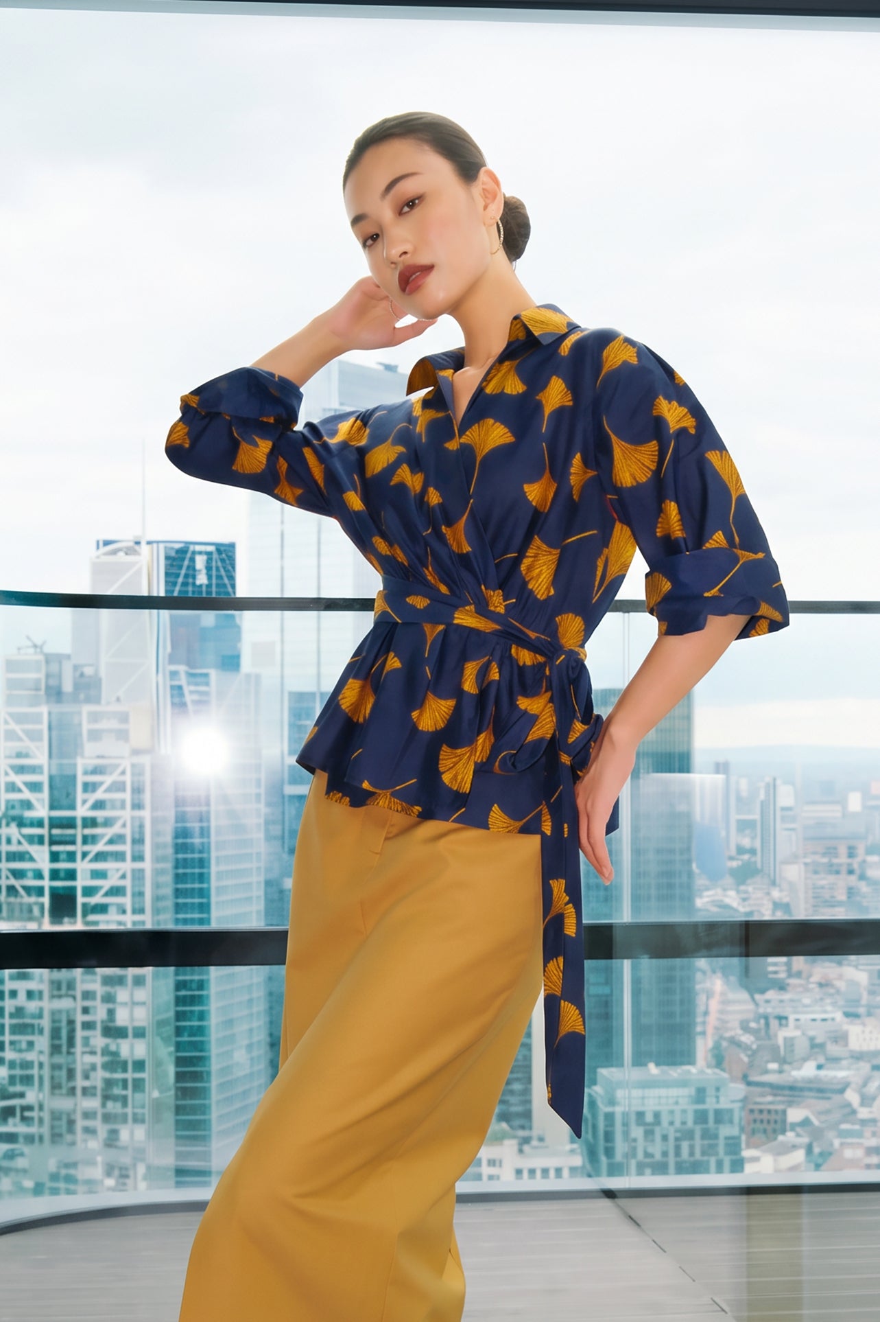 A stylish woman posing against a city skyline wearing a navy blue wrap blouse with golden ginkgo leaf print tied at the waist, paired with tailored mustard yellow wide-leg trousers.