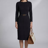 A professional woman wearing a sophisticated black long-sleeve sheath dress, featuring a classic crew neckline and a slim black belt with a gold buckle for a polished executive office look.