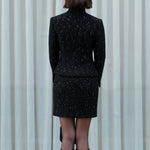 A detailed shot of a woman modeling a sophisticated black woven tweed mini skirt adorned with subtle shimmering crystals, tailored in a sleek high-waisted pencil cut.