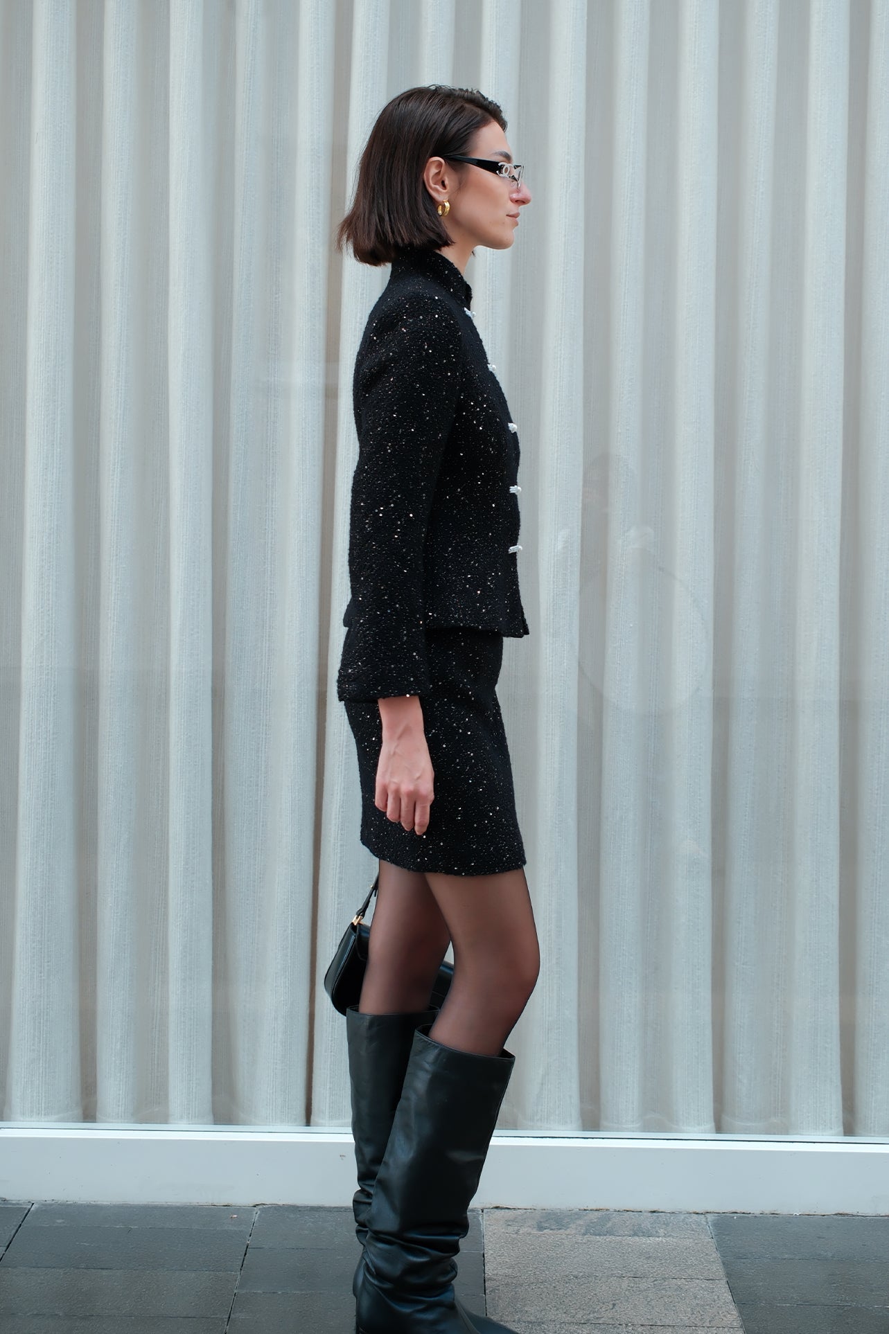 A chic woman wearing a luxurious black sparkle tweed tailored jacket featuring an authentic stand cheongsam collar, elegant horizontal pearl bar closures, and a flattering hourglass waistline.