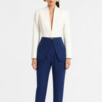 A model showcasing elegant navy blue high-waisted work trousers, featuring a unique curved overlay panel design at the waist, a slim golden metal bar buckle, pressed front creases, and chic cuffed hems, paired with a light blue button-up shirt.