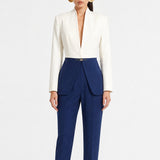 A model showcasing elegant navy blue high-waisted work trousers, featuring a unique curved overlay panel design at the waist, a slim golden metal bar buckle, pressed front creases, and chic cuffed hems, paired with a light blue button-up shirt.