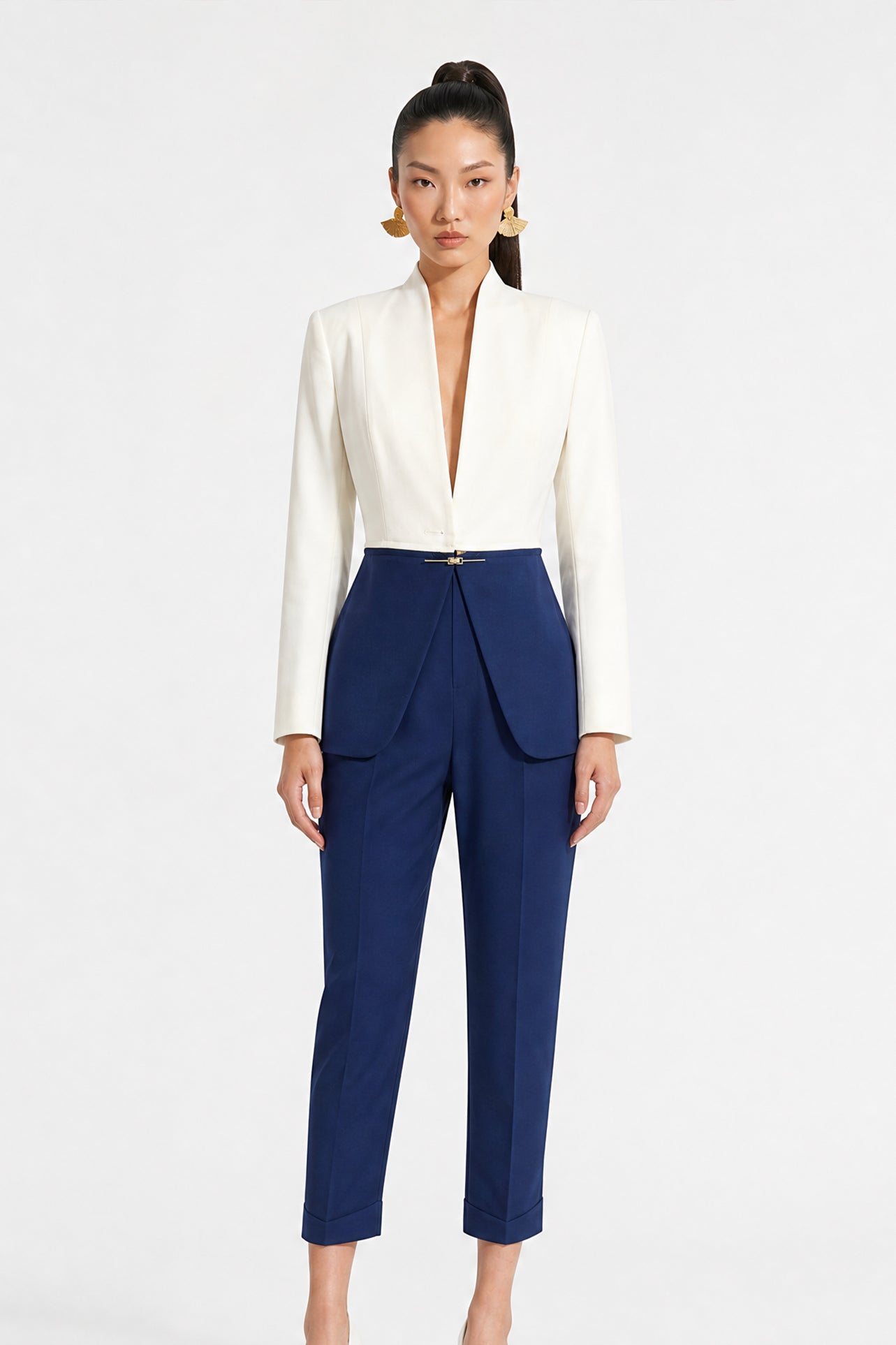A model showcasing elegant navy blue high-waisted work trousers, featuring a unique curved overlay panel design at the waist, a slim golden metal bar buckle, pressed front creases, and chic cuffed hems, paired with a light blue button-up shirt.