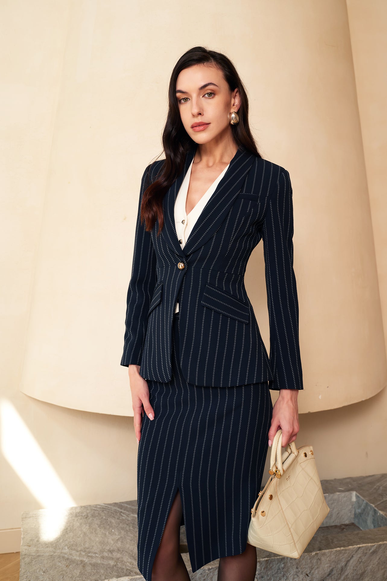 A professional woman wearing a sophisticated navy blue pinstripe suit, featuring a tailored single-button blazer and a matching midi pencil skirt with a front slit for an elegant executive office look.