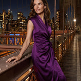A professional woman modeling an elegant deep purple satin wrap midi dress, featuring a sophisticated surplice V-neck, cap sleeves, and a chic side bow tie for a quiet luxury evening or office look.