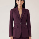  A professional woman wearing glasses and an elegant deep burgundy skirt suit featuring a sculptural stand-up lapel blazer with a single-button closure, tailored flap pockets, and a matching high-waisted pencil skirt.