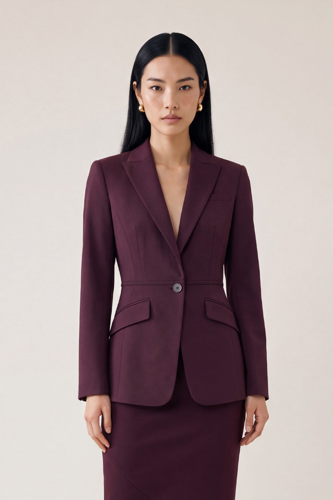  A professional woman wearing glasses and an elegant deep burgundy skirt suit featuring a sculptural stand-up lapel blazer with a single-button closure, tailored flap pockets, and a matching high-waisted pencil skirt.