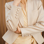 A professional woman modeling a luxurious champagne satin blazer featuring a soft shawl lapel and a single-button closure, elegantly paired with high-waisted white trousers for a sophisticated quiet luxury look.