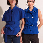 A style comparison of two professional women wearing modern cobalt blue outerwear featuring self-tie waist belts; on the left, a tailored cropped capelet jacket paired with jeans, and on the right, a double-breasted sleeveless vest with white contrast buttons styled with black trousers.