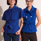 A style comparison of two professional women wearing modern cobalt blue outerwear featuring self-tie waist belts; on the left, a tailored cropped capelet jacket paired with jeans, and on the right, a double-breasted sleeveless vest with white contrast buttons styled with black trousers.