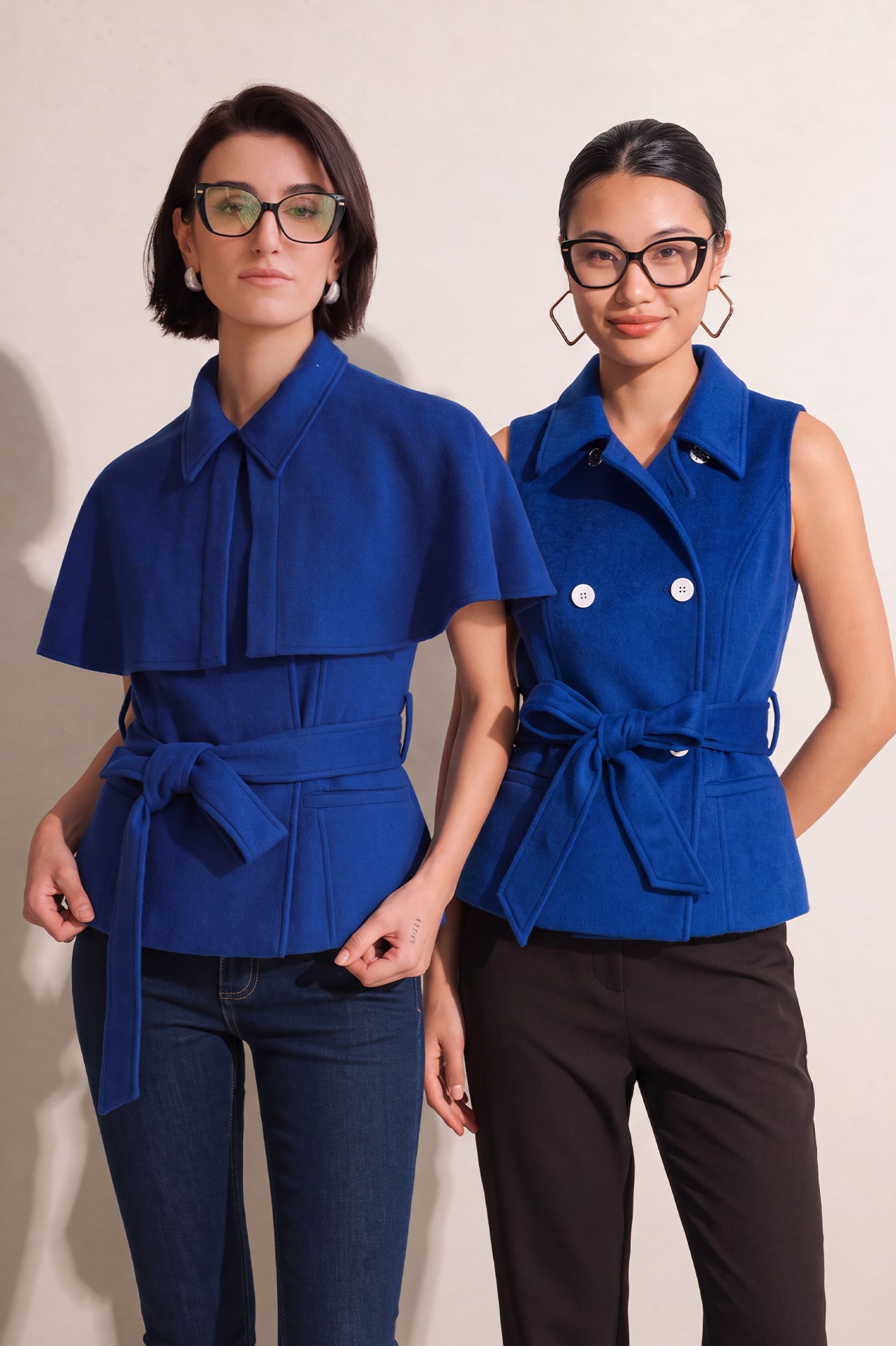 A style comparison of two professional women wearing modern cobalt blue outerwear featuring self-tie waist belts; on the left, a tailored cropped capelet jacket paired with jeans, and on the right, a double-breasted sleeveless vest with white contrast buttons styled with black trousers.
