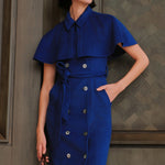 An elegant woman wearing a sophisticated royal cobalt blue trench dress featuring a structured turn-down collar, a cropped capelet overlay, a double-breasted button front, and a cinched self-tie belted waist.