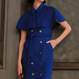 An elegant woman wearing a sophisticated royal cobalt blue trench dress featuring a structured turn-down collar, a cropped capelet overlay, a double-breasted button front, and a cinched self-tie belted waist.