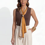A professional woman modeling a sleeveless chocolate brown top and cream wide-leg pleated trousers, styled with a patterned yellow silk scarf and bold gold jewelry for a sophisticated office aesthetic.