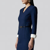 A professional woman wearing a sophisticated navy blue V-neck sheath dress, featuring elegant white contrast turn-up cuffs and a slim black belt with a gold buckle for a polished executive look.