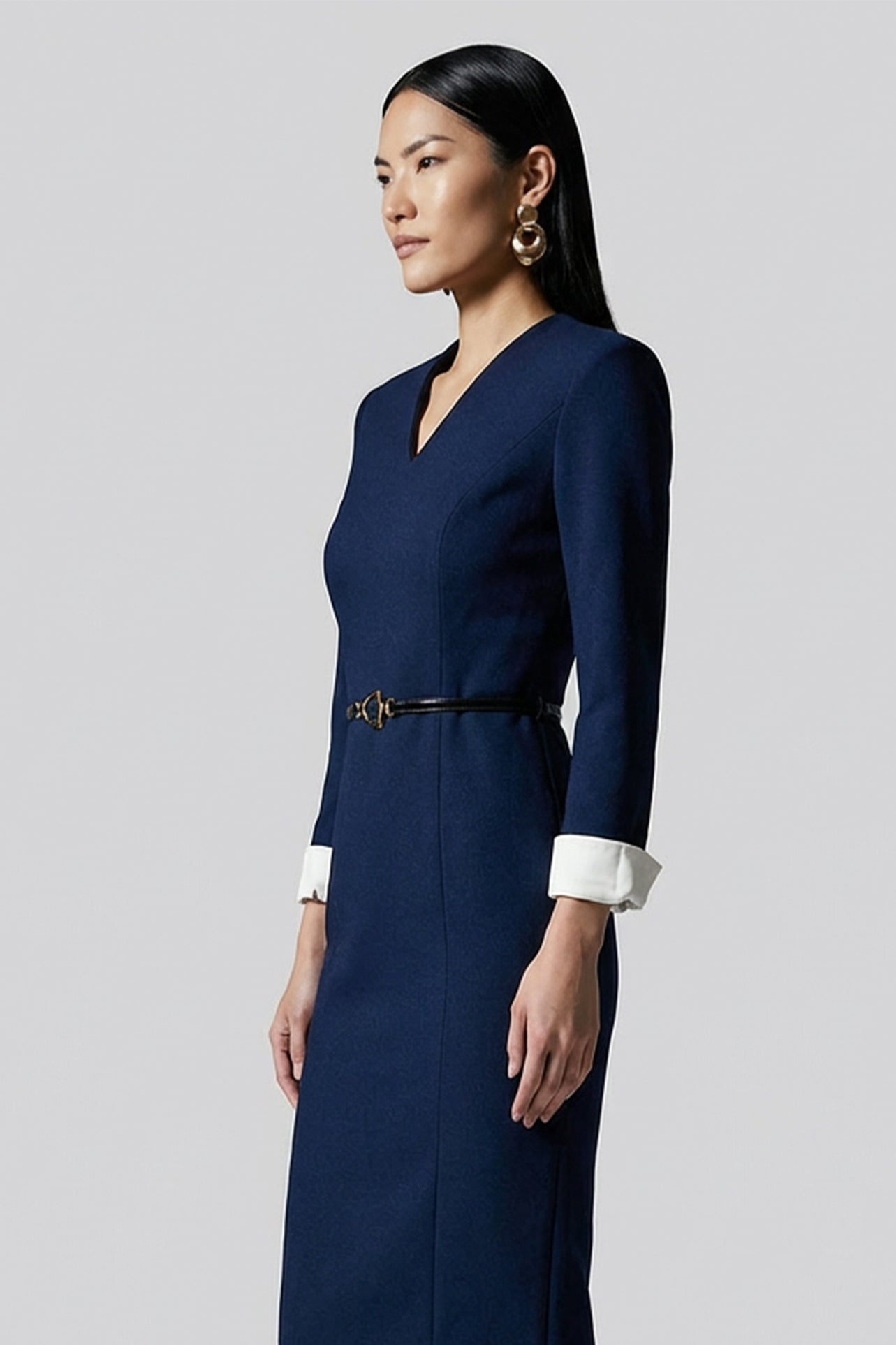 A professional woman wearing a sophisticated navy blue V-neck sheath dress, featuring elegant white contrast turn-up cuffs and a slim black belt with a gold buckle for a polished executive look.