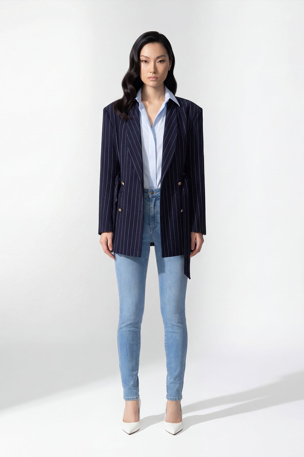 A professional woman wearing a sophisticated navy blue pinstripe blazer jacket, featuring a longline silhouette with a matching tie-waist belt and gold earring accents for an elegant executive office look.