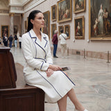 An elegant professional woman wearing a sophisticated white double-breasted coat dress featuring sharp navy blue contrast piping and a matching waist belt for a tailored executive look